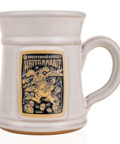 The front of the Bones Coffee Company White Rabbit Handthrown Mug. It has the art for White Rabbit on the golden medallion. The mug is white colored.