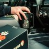 The black 40 ounce tumbler is in a cupholder in a cart. Someone's hand is on top of the lid and the box the 40 ounce tumbler it came in is in the passenger seat of the car.
