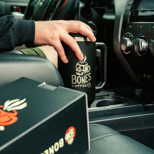 The black 40 ounce tumbler is in a cupholder in a cart. Someone's hand is on top of the lid and the box the 40 ounce tumbler it came in is in the passenger seat of the car.