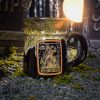 The front of the Bones Coffee Company Army of Dark Chocolate Handthrown Mug. It has the Army of Dark Chocolate art on the golden medallion. The mug is gloss black colored with a sunshine and white glaze. The mug is in a graveyard on top of gravel.