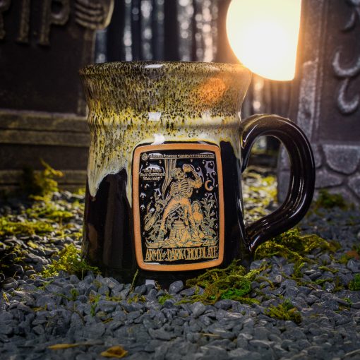 The front of the Bones Coffee Company Army of Dark Chocolate Handthrown Mug. It has the Army of Dark Chocolate art on the golden medallion. The mug is gloss black colored with a sunshine and white glaze. The mug is in a graveyard on top of gravel.