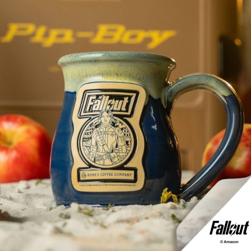 The Lucy hand thrown mug is sitting in sand with a pip-boy case and apples nearby. The mug is colored powder blue with a yellow and white glaze on top, and has the Atomic Apple art on the golden medallion.
