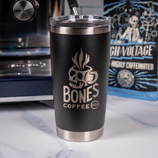 A black tumbler is on top of a marble surface with an espresso machine and High Voltage 12 ounce bag behind it. On the tumbler is the Skull Logo for Bones Coffee Company that has a skull that looks like a mug on it.