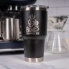 A black tumbler is on top of a marble surface with an espresso machine and coffee dripper behind it. On the tumbler is the Skull Logo for Bones Coffee Company that has a skull that looks like a mug on it.