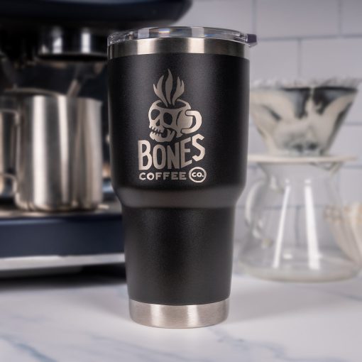 A black tumbler is on top of a marble surface with an espresso machine and coffee dripper behind it. On the tumbler is the Skull Logo for Bones Coffee Company that has a skull that looks like a mug on it.