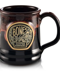 The front of the Bones Coffee Company Classic Logo mug that is black colored with an orange glaze on top of it. There is a gold medallion on the center of the mug that has the classic logo for Bones Coffee Company of a skeleton sipping coffee on top of greenery on it.