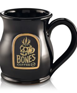 The front of the Bones Coffee Company Skull Logo mug that is black colored. There is a gold medallion on the center of the mug that has the skull logo for Bones Coffee Company of a skull that looks like a mug with steam on top of it on it.