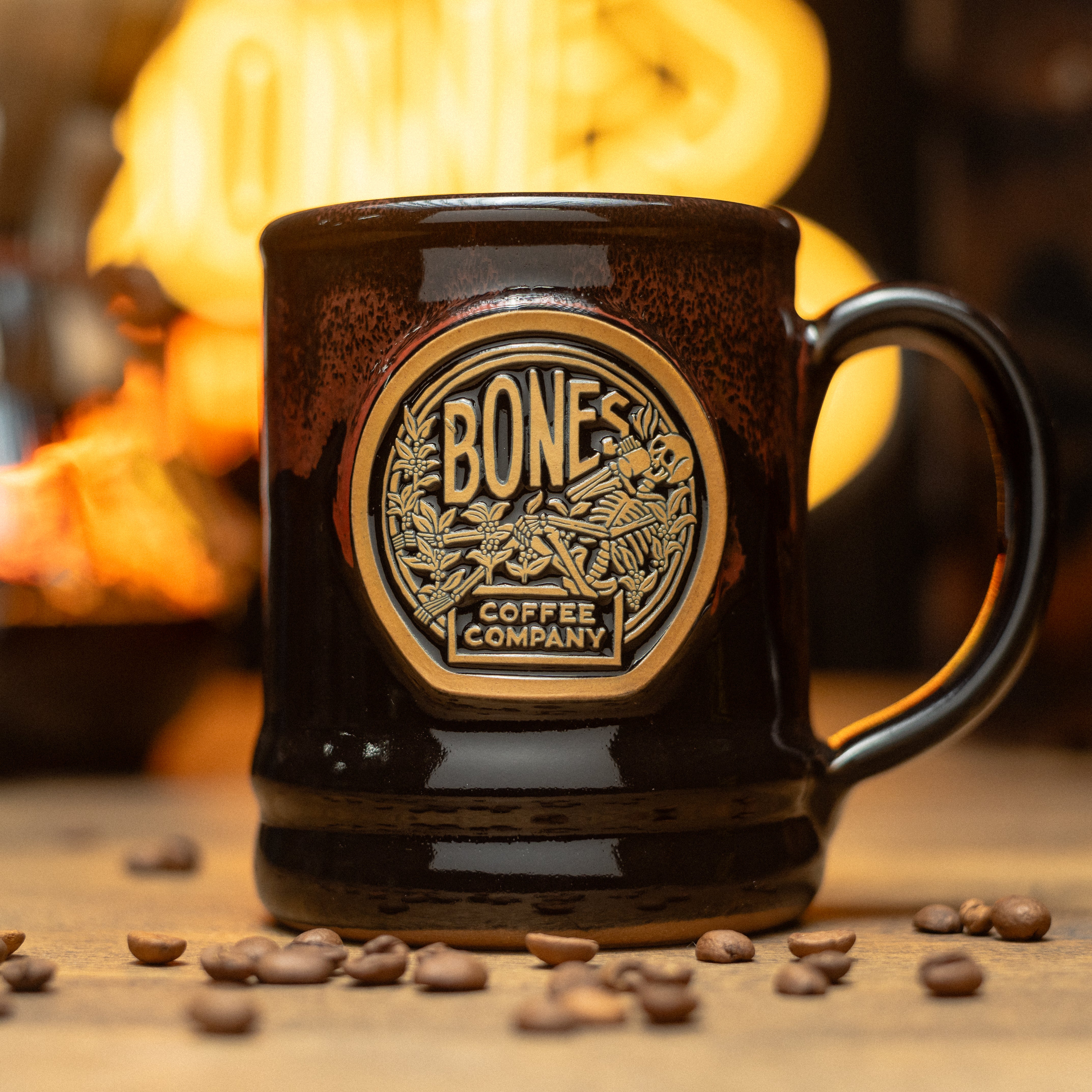 The Classic Logo handthrown mug is on top of a brown surface with a neon sign behind it and coffee beans nearby it. The mug is black colored with an orange glaze on top of it. The golden medallion on it has the Classic Logo of Bones Coffee Company on it.