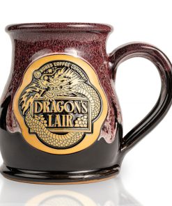 The front of the Bones Coffee Company Dragon’s Lair hand thrown mug with the Dragon’s Lair art on the golden medallion. The mug is black colored with a burgundy and white glaze on top.
