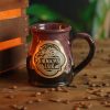 The Dragon’s Lair mug is on a wooden table next to a glowing wooden crate. Coffee beans are scattered around the surface. The mug is black colored with a burgundy and white glaze on top of it with a golden medallion on the front that shows the Dragon’s Lair art on it.