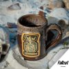 The Ghoul hand thrown mug is sitting in sand and rubble with a skull nearby. The mug is colored chocolate with a sand-white glaze on top, and it has the Wasteland Crunch art on the golden medallion.