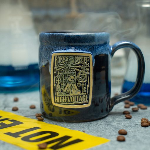 The High Voltage handthrown mug is on top of a rocky surface with beakers behind it and coffee beans and caution tape nearby it. The mug is black colored with a blue glaze on top of it. The golden medallion on it has the High Voltage coffee art on it.