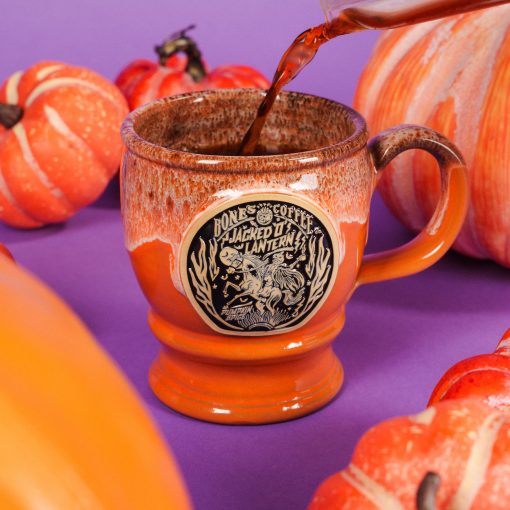 The front of the Bones Coffee Company Jacked O’ Lantern hand thrown mug. The golden medallion has the art of the Jacked O’ Lantern coffee on it. The mug is an orange finish dipped in pumpkin spice glaze. There are pumpkins around it and coffee is being poured inside.