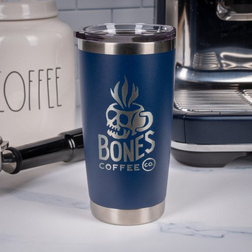 A navy blue tumbler is on top of a marble surface with an espresso machine and coffee container behind it. On the tumbler is the Skull Logo for Bones Coffee Company that has a skull that looks like a mug on it.