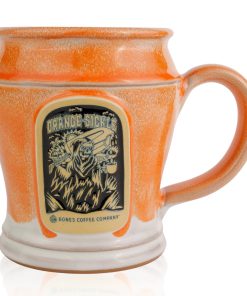The front of the Bones Coffee Company Orange-Sickle hand thrown mug with the Orange-Sickle coffee art on the golden medallion. The mug is white colored with an orange glaze on top.