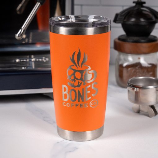 An orange tumbler is on top of a marble surface with an espresso machine and coffee grinder behind it. On the tumbler is the Skull Logo for Bones Coffee Company that has a skull that looks like a mug on it.