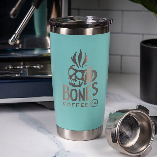 A robin’s egg blue tumbler is on top of a marble surface with an espresso machine and cup behind it. On the tumbler is the Skull Logo for Bones Coffee Company that has a skull that looks like a mug on it.