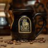 The Skull Logo handthrown mug is on top of a brown surface with a coffee pot behind it and coffee beans nearby it. The mug is black colored. The golden medallion on it has the Skull Logo of Bones Coffee Company on it.