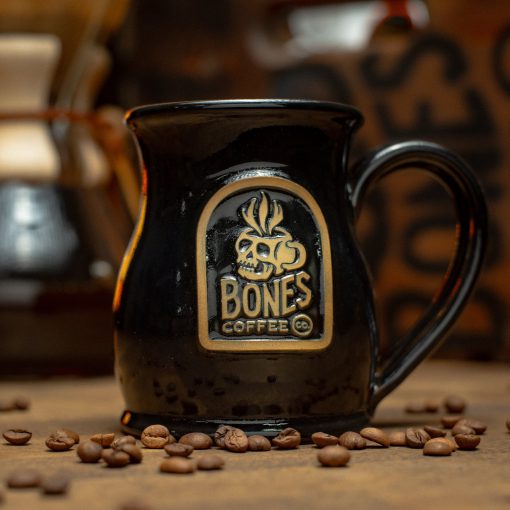The Skull Logo handthrown mug is on top of a brown surface with a coffee pot behind it and coffee beans nearby it. The mug is black colored. The golden medallion on it has the Skull Logo of Bones Coffee Company on it.