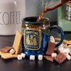 The front of the Bones Coffee Company S’morey Time Handthrown Mug. It has the S’morey Time art on the golden medallion. The mug is blue colored with a cinnamon and white glaze. The mug is on a black surface with chocolate, marshmallows, and graham crackers nearby.