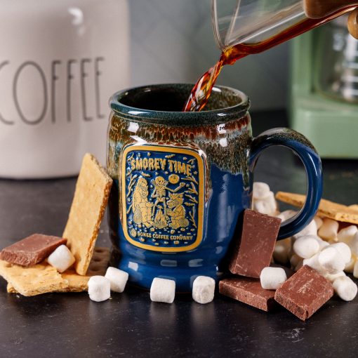 The front of the Bones Coffee Company S’morey Time Handthrown Mug. It has the S’morey Time art on the golden medallion. The mug is blue colored with a cinnamon and white glaze. The mug is on a black surface with chocolate, marshmallows, and graham crackers nearby.