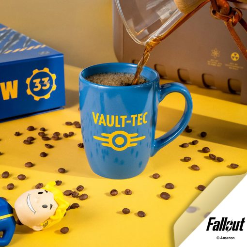 The Vault-Tec ceramic mug is colored blue with a yellow logo of Vault-Tec on it. Coffee is being poured into it. A Vault Boy, Fallout Collector’s Box, Pip-Boy case, and coffee beans are nearby.