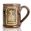 The front of the Bones Coffee Company Ghoul hand thrown mug with the Wasteland Crunch art on the golden medallion. It is inspired by Zenimax and Amazon’s Fallout show. The mug is chocolate colored with a sand white glaze on top.