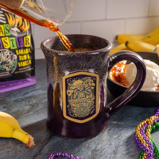 The front of the Bones Coffee Company Bananas Foster Handthrown Mug. It has the art for Bananas Foster coffee on the golden medallion. The mug is a deep purple color and has a banana and white glaze on top of it. There is a bananas foster, bananas, and beads nearby the mug on a black surface.