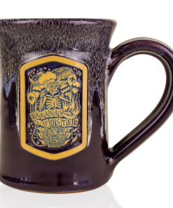 The front of the Bones Coffee Company Bananas Foster Handthrown Mug. It has the art for Bananas Foster coffee on the golden medallion. The mug is a deep purple color and has a banana and white glaze on top of it.