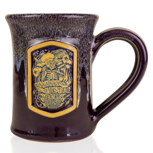 The front of the Bones Coffee Company Bananas Foster Handthrown Mug. It has the art for Bananas Foster coffee on the golden medallion. The mug is a deep purple color and has a banana and white glaze on top of it.