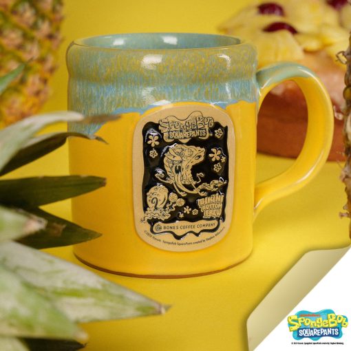 The front of the Bones Coffee Company Bikini Bottom Brew hand thrown mug with SpongeBob SquarePants and Gary on the golden medallion. It is inspired by SpongeBob Squarepants. The mug is yellow colored and has a blue and white glaze on top of it. It has pineapples nearby.