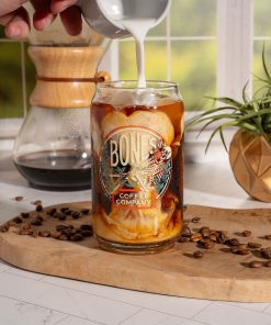 A cold brew coffee glass that has the Bones Coffee Company logo on it. The skeleton in the logo is sipping coffee and relaxing on greenery. Coffee and ice is inside the glass and someone is pouring cream into it.