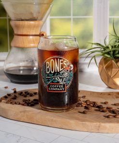 A cold brew coffee glass that has the Bones Coffee Company logo on it. The skeleton in the logo is sipping coffee and relaxing on greenery. Coffee and ice is inside the glass.