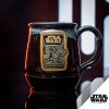 The Dark Side Chocolate Truffle hand thrown mug is in front of a metal panel. The metal panel has light coming through the rounded rectangles. The mug is colored black with a white glaze on top of it, and the gold medallion on the mug has the Dark Side Chocolate Truffle art on it.