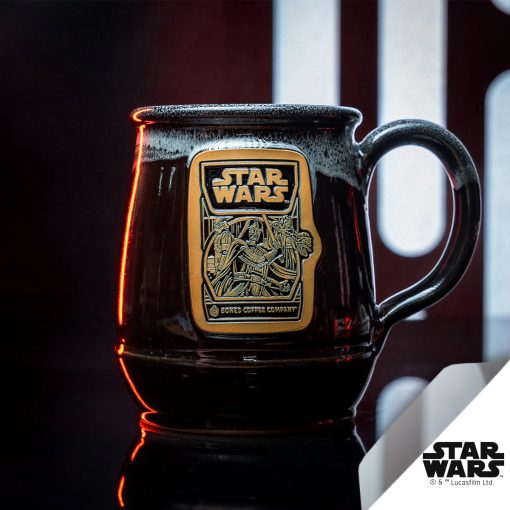 The Dark Side Chocolate Truffle hand thrown mug is in front of a metal panel. The metal panel has light coming through the rounded rectangles. The mug is colored black with a white glaze on top of it, and the gold medallion on the mug has the Dark Side Chocolate Truffle art on it.