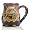 The front of the Bones Coffee Company Dragon’s Lair hand thrown mug with the Dragon’s Lair art on the golden medallion. The mug is black colored with a burgundy and white glaze on top.