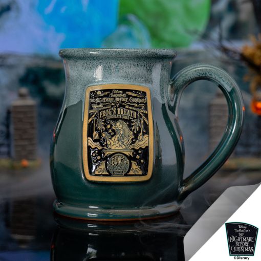 The front of the Bones Coffee Company Frog's Breath hand thrown mug with Sally on the golden medallion. It is inspired by Disney Tim Burton’s The Nightmare Before Christmas. The mug is green colored with a white glaze on top of it. It is inside a foggy toy graveyard.