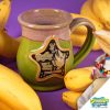 The front of the Bones Coffee Company Goofy Goober Sundae hand thrown mug with Patrick and a jellyfish on the golden medallion. It is inspired by SpongeBob Squarepants. The mug is moss green colored and has a pink and white glaze on top of it. It has bananas and a banana split sundae nearby.