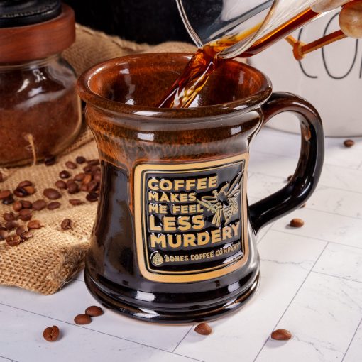The front of the Bones Coffee Company Murder Hornet Handthrown Mug. It has a hornet with a knife and it says coffee makes me feel less murdery on the golden medallion. The mug is black colored with a sunset orange glaze. There are coffee beans near the mug on a marble surface.