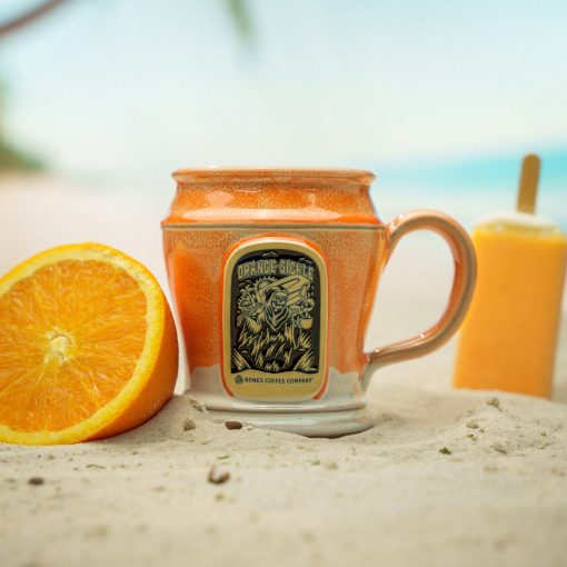 The front of the Bones Coffee Company Orange-Sickle hand thrown mug with the Orange-Sickle coffee art on the golden medallion. The mug is white colored with an orange glaze on top. It is on the beach with an orange and orange creamsicle nearby.