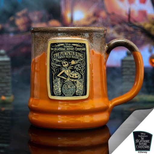 The front of the Bones Coffee Company The Pumpkin King hand thrown mug with Jack Skellington on the golden medallion. It is inspired by Disney Tim Burton’s The Nightmare Before Christmas. The mug is orange colored with a black and white glaze on top. It is inside a toy graveyard.