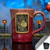 The front of the Bones Coffee Company Santa Jack hand thrown mug with Jack Skellington on the golden medallion. It is inspired by Disney Tim Burton’s The Nightmare Before Christmas. The mug is cranberry colored with a white glaze on top. It is inside a toy graveyard.