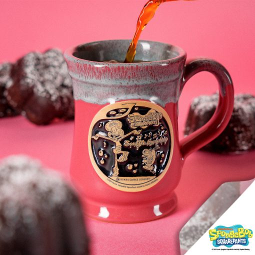 The front of the Bones Coffee Company Souffle Symphony hand thrown mug with Squidward and Plankton on the golden medallion. It is inspired by SpongeBob Squarepants. The mug is cosmo pink colored and has a teal and white glaze on top of it. It has chocolate souffles nearby.