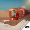 The Twin Sun Tiramisu hand thrown mug is on a rock surrounded by sand. The mug is paprika colored with a yellow glaze on top, and it has the Twin Sun Tiramisu coffee art on a golden medallion.