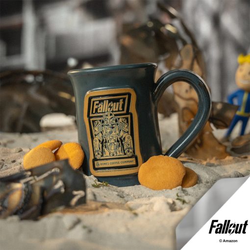 The Maximus hand thrown mug is sitting in sand and rubble with a Vault Boy and vanilla wafers nearby. The mug is colored gray and has the Valiant Vanilla art on the golden medallion.
