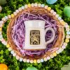 The front of the Bones Coffee Company White Rabbit hand thrown mug, and it has the White Rabbit art on the golden medallion. The mug is white colored. It is in a basket with pink plastic strips under it, and there are easter eggs around the basket.
