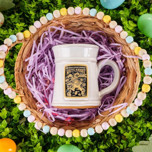 The front of the Bones Coffee Company White Rabbit hand thrown mug, and it has the White Rabbit art on the golden medallion. The mug is white colored. It is in a basket with pink plastic strips under it, and there are easter eggs around the basket.
