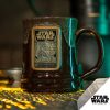The Wookiee Cookie hand thrown mug is set atop a grate with light beneath it. There are metal parts in the background. The mug is chocolate colored with a sand-white glaze on top of it. Its golden medallion features the art from the Wookiee Cookie coffee.